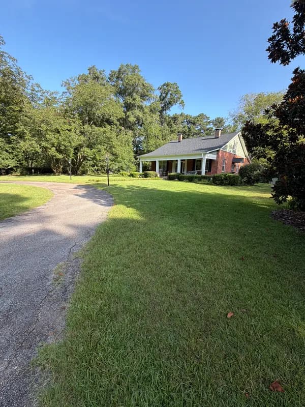 Brick home with white columned porch behind a green lawn lined by a curved driveway and mature trees