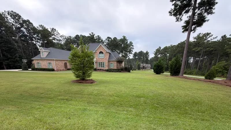 Wide green lawn with a young tree and large brick two-story home under a cloudy sky
