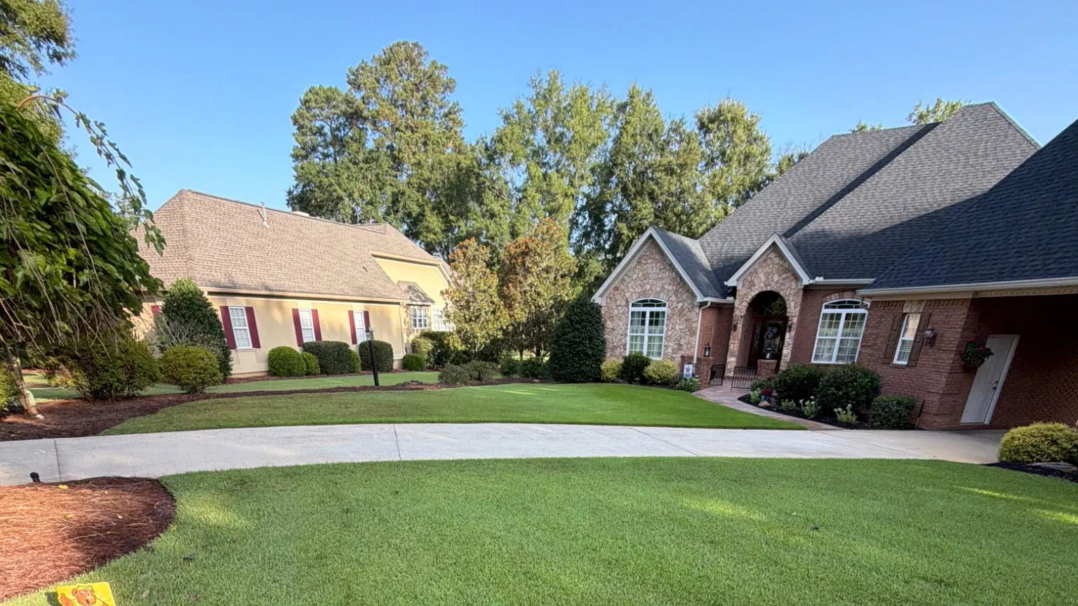 Lush green lawn leading to a brick and stone home with mature trees under a clear blue sky.