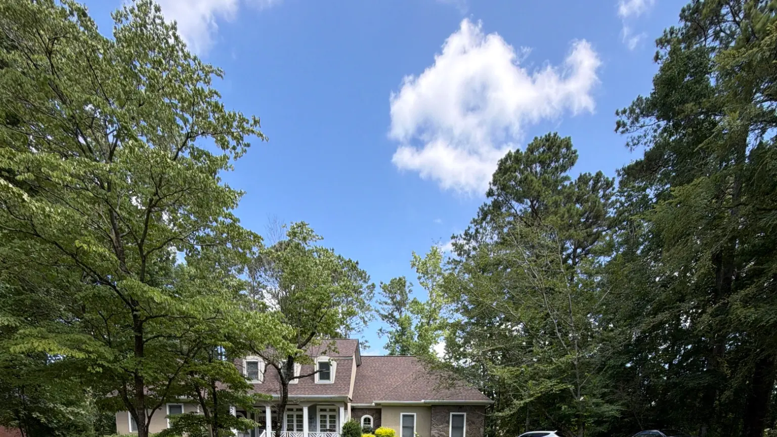 Wide green lawn leading up to a two-story house with a covered porch and driveway, framed by trees