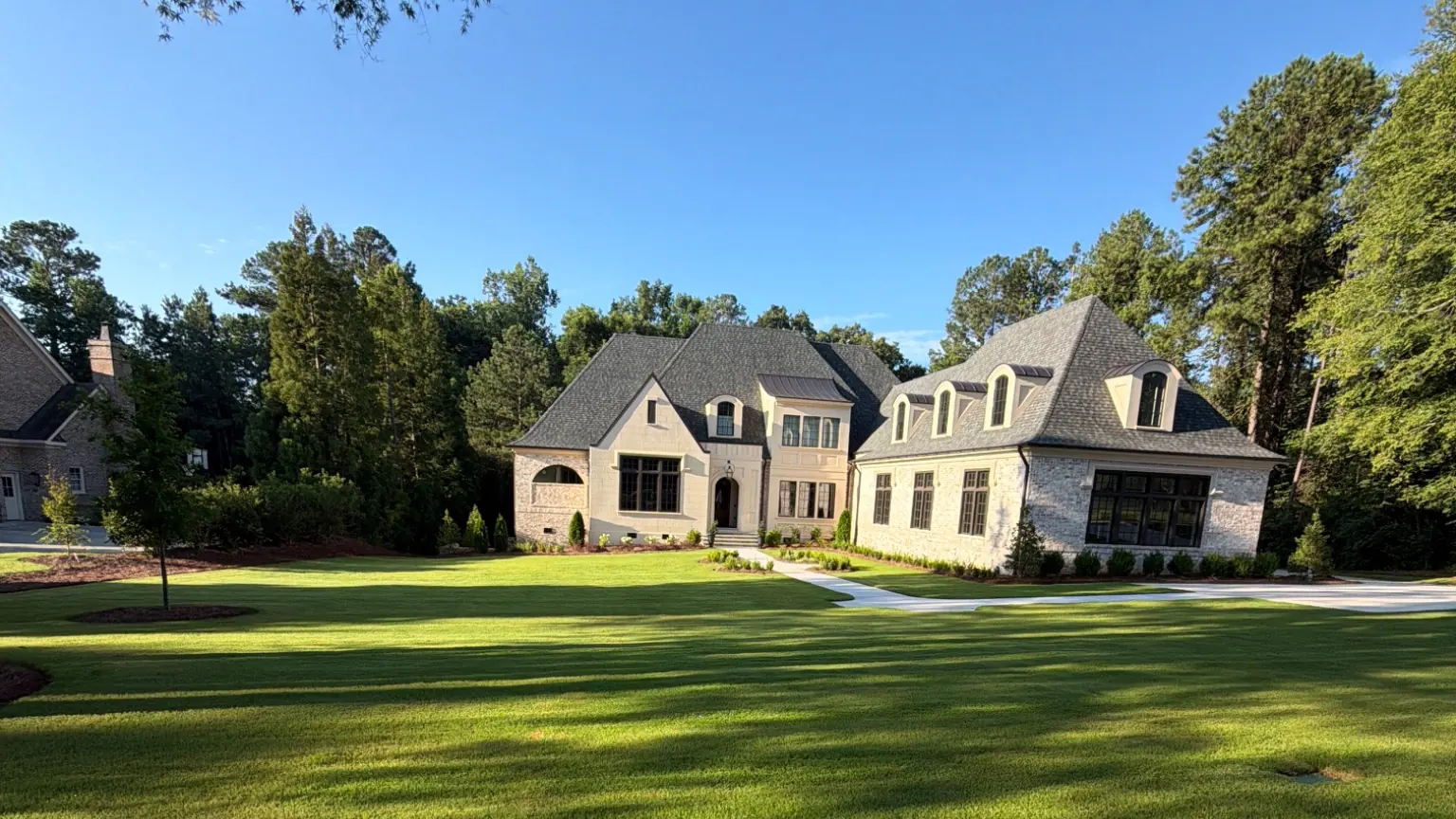 Large stone home with lush green lawn, landscaped beds, and mature trees under blue sky